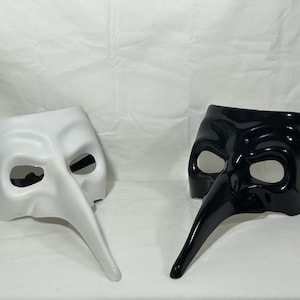 May include: Two Venetian masks, one white and one black, are displayed against a white backdrop. Each mask features a long, curved beak and eye openings. The masks are likely for costume or decorative purposes.