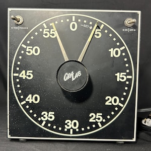 May include: A square black GraLab darkroom timer with a large circular face featuring white numbers from 0 to 55. It has two metal toggle switches at the top corners labeled Time, Focus, On, and Off, and a central black knob with the GraLab logo.