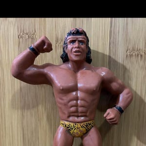 May include: A plastic action figure of a wrestler with a muscular build. The figure is wearing a yellow and black leopard print loincloth. The figure has brown skin and black hair.