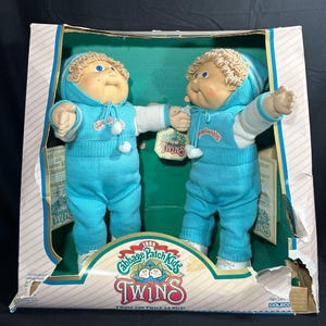 May include: A 1985 Cabbage Patch Kids Twins doll set in original packaging. Two dolls with blonde yarn hair, blue eyes, and light skin wear matching bright blue knit hooded outfits with white accents. The box features the Cabbage Patch Kids logo.