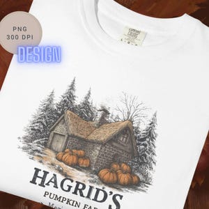 May include: A white t-shirt featuring a graphic illustration of a stone cottage surrounded by pine trees and orange pumpkins. The text below the illustration reads HAGRID'S PUMPKIN FARM. The shirt has a Comfort Colors brand label at the neck.