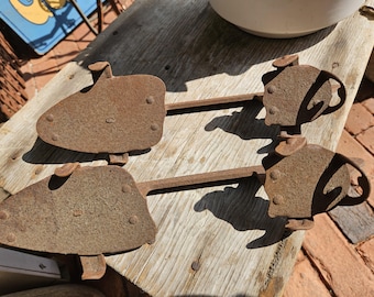 May include: Two antique, rusty metal shoe stretchers. Each stretcher features a shoe-shaped plate with rivets, linked to a handle and a heel piece. The items are displayed on a weathered wooden surface.