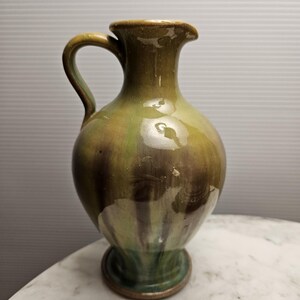 May include: A green and brown ceramic jug with a wide base and a handle. The jug has a glossy finish and a unique glaze pattern.