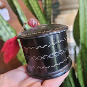 Puede incluir: Un pequeño recipiente cilíndrico negro con un patrón plateado decorativo alrededor del cuerpo. Una borla roja y un pequeño adorno de cuentas rojas cuelgan de la parte superior. El recipiente se sostiene en una mano.