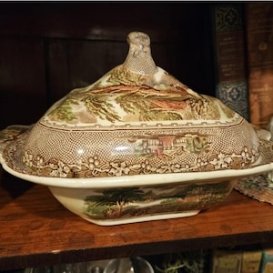 May include: A vintage ceramic serving dish with a lid, featuring a detailed landscape design in shades of brown, green, and beige. The dish has a decorative rim and a textured pattern, ideal for serving food. A classic piece for any kitchen.