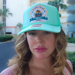 Pode incluir: Um boné de caminhoneiro turquesa com uma malha branca nas costas. O boné tem um remendo na frente que diz "Panama City Beach, Flórida" com uma palmeira e uma cena de praia. O texto "Everything" está em um círculo ao redor do remendo.