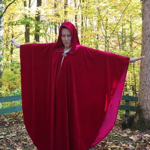 May include: A long, red velvet cape with a hood. The cape is being worn by a person standing in a woodland setting.