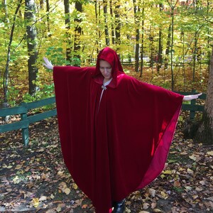May include: A woman wearing a long, red velvet cape with a hood. The cape is flowing in the wind and the woman is standing in a woodland with autumn foliage.
