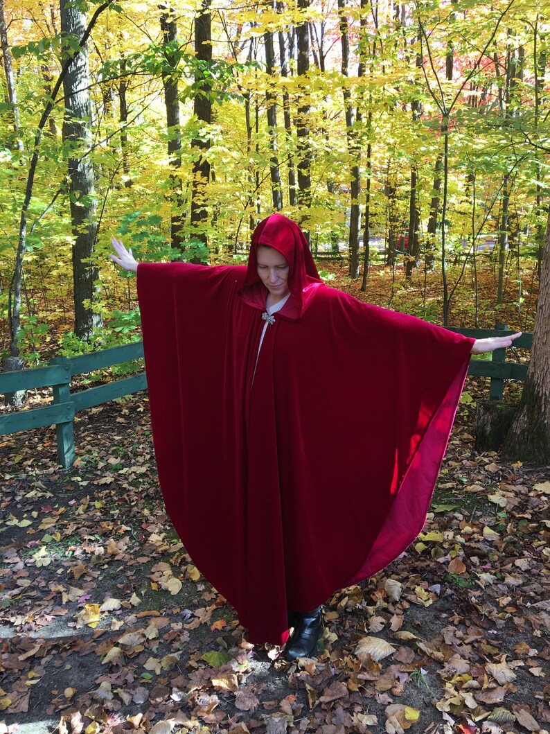 May include: A woman wearing a long, red velvet cape with a hood. The cape is flowing in the wind and the woman is standing in a woodland with autumn foliage.