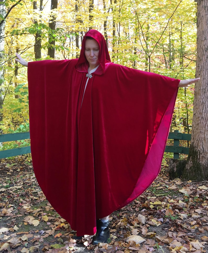 May include: A long, red velvet cape with a hood. The cape is being worn by a person standing in a woodland setting.