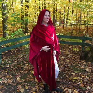 May include: A woman wearing a long, red velvet hooded cape in a woodland setting. The cape has a flowing design and is draped over her shoulders. The woodland is filled with fallen leaves in shades of red, orange, and yellow.