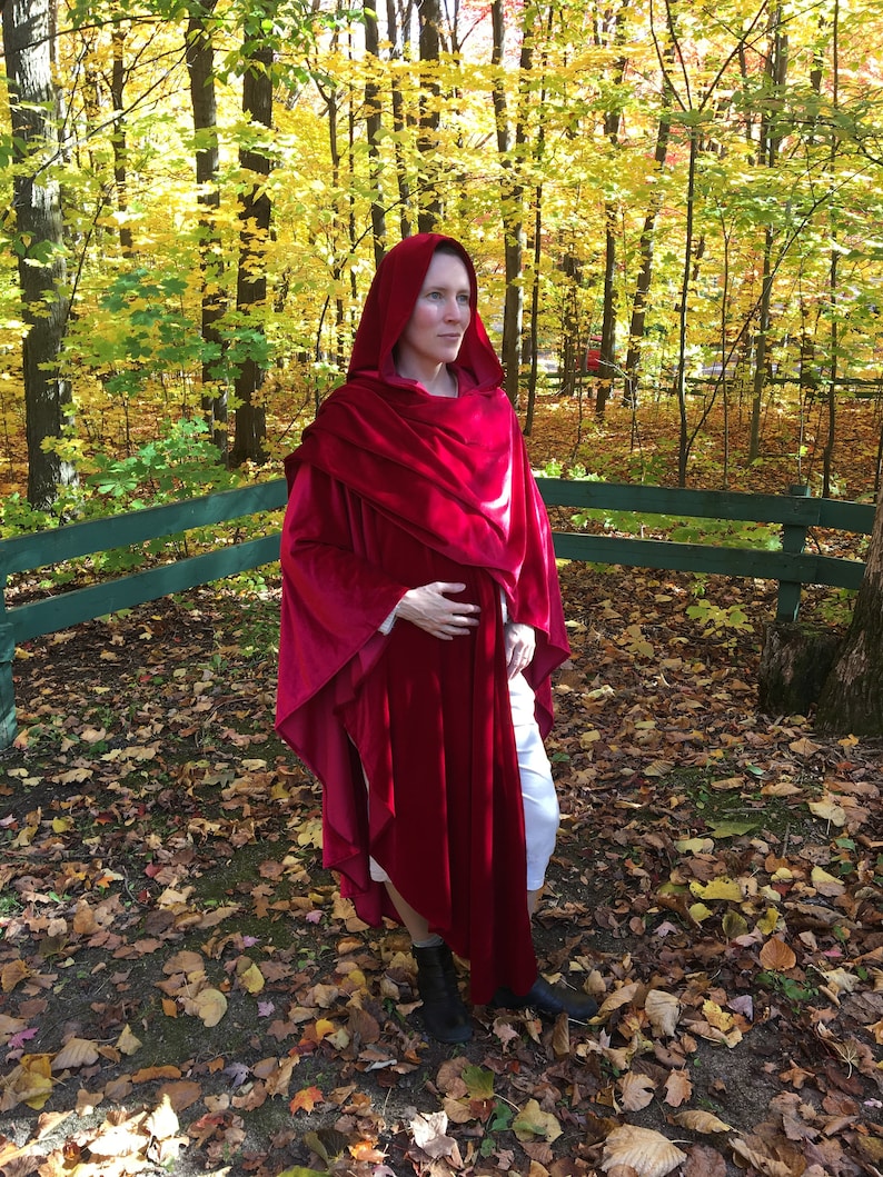 May include: A woman wearing a long, red velvet hooded cape in a woodland setting. The cape has a flowing design and is draped over her shoulders. The woodland is filled with fallen leaves in shades of red, orange, and yellow.