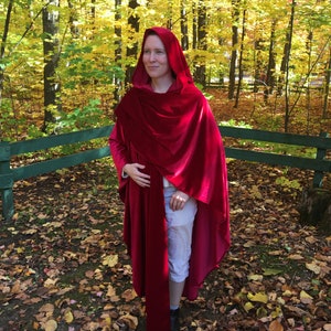 May include: A woman wearing a long, red velvet hooded cape in a woodland setting. The cape is draped over her shoulders and has a long train. The woman is standing in a forest with fallen leaves on the ground.