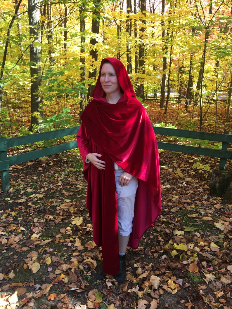 May include: A woman wearing a long, red velvet hooded cape in a woodland setting. The cape is draped over her shoulders and has a long train. The woman is standing in a forest with fallen leaves on the ground.