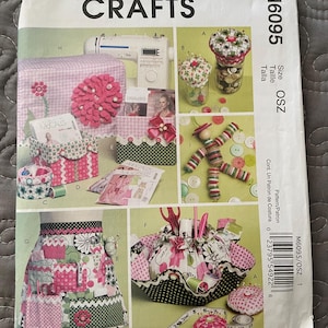 May include: A McCall's Crafts sewing pattern package featuring various craft projects. The package includes images of sewing projects like a sewing machine cover, pincushions, a sewing basket, and an apron. The color scheme is primarily pink, green, and white.