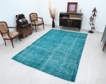 May include: A large, rectangular turquoise rug with a distressed, vintage appearance. The rug is the focal point of a room with antique furniture, including chairs, a cabinet, and a wooden chest. The room has a neutral color palette with white walls and light-colored flooring.