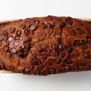 May include: A rectangular loaf of baked banana bread topped with numerous dark brown chocolate chips. The bread is contained within a light tan paper baking liner, showcasing a textured, golden-brown surface with a visible crack down the centre.
