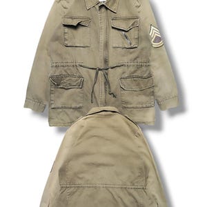 Pode incluir: Uma jaqueta de campo verde-oliva com gola, dragonas abotoadas, quatro bolsos frontais e cintura com cordão. Um emblema de chevron de estilo militar está na manga esquerda. O texto diz: CHEST 20, LENGTH 28.5.