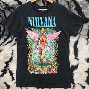 Puede incluir: Camiseta negra con la palabra "NIRVANA" en turquesa sobre un gráfico. El gráfico representa una figura con alas rosas, rodeada de una escena floral y forestal. La camiseta está colgada en una percha de madera.