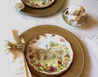 May include: A table setting featuring gold-colored chargers, floral patterned plates, a teacup and saucer, and folded napkins with gold bow napkin rings. The plates display a scenic landscape with flowers. The setting is on a white lace tablecloth.