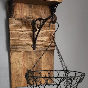 May include: A rustic wooden wall shelf with a hanging planter. The shelf has a small top shelf with two small ceramic pots, one red and one blue. The hanging planter is a black metal basket with a decorative design, suspended from a chain attached to a black metal hook.