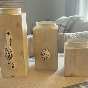 May include: Three white wooden candle holders with a distressed finish. The candle holders have a simple design with a round top and a square base. The candle holders are arranged in a row on a table.