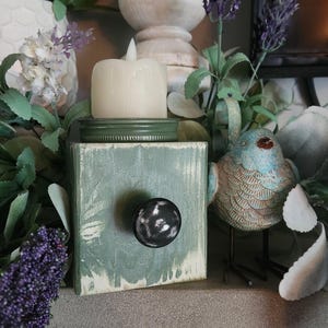 May include: A green wooden box with a black knob and a white candle inside. The box is sitting on a stone surface with a blue bird figurine and green and white plants.