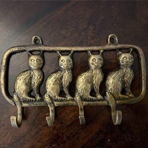 Könnte beinhalten: Schlüsselhalter aus Messing mit vier Katzen-Silhouetten und Haken. Die Katzen stehen in einer Reihe, nach vorne gerichtet, mit ihren Schwänzen um die Haken gewickelt. Der Schlüsselhalter ist rechteckig mit einem dekorativen Rand.