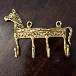 May include: Gold metal horse-shaped key holder with five hooks. The horse has intricate details and a patterned design.