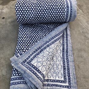 Op de afbeelding: Een opgerolde marineblauwe en witte quilt met een geometrisch patroon. De quilt is gedeeltelijk uitgerold en onthult een wit gedeelte met een bloemenontwerp en een marineblauwe rand. De quilt is gemaakt van een zacht materiaal.