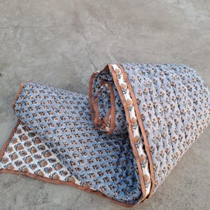 Op de afbeelding: Een opgerolde, lichtblauwe quilt met een herhalend bloemmotief in bruin en wit. De quilt heeft een bruine rand en is gemaakt van een zachte, gewatteerde stof. Het ontwerp is symmetrisch.