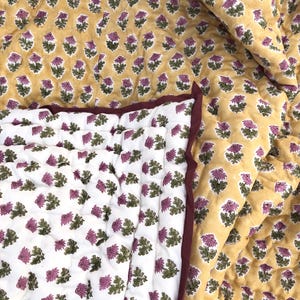 May include: A quilted blanket with a floral pattern. The top side is mustard yellow with purple and green flowers. The underside is white with the same floral design. The edges are trimmed with a dark burgundy fabric.