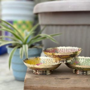 Op de afbeelding: Drie decoratieve messing kommen met sierlijke randen en bloemmotieven. De kommen zijn gestapeld op een houten oppervlak. De kommen hebben een diameter van ongeveer 10 cm.