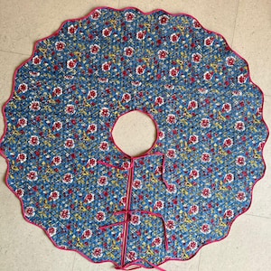 Op de afbeelding: Een blauwe gewatteerde boomrok met een bloemenpatroon van rode en gele bloemen. De rok heeft een geschulpte rand met een roze bies en strikken. Het midden heeft een cirkelvormige opening voor de stam.