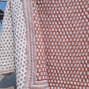 May include: A quilted blanket with a floral pattern in shades of white and brown. The blanket has a detailed border and a contrasting pattern on the reverse side. The fabric appears soft and comfortable, suitable for a bed or sofa.