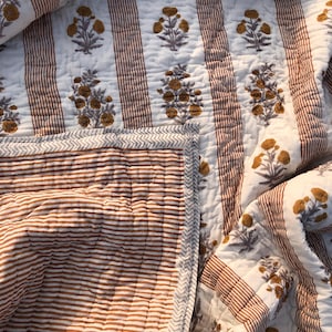 May include: A white and brown floral quilt with a striped pattern. The quilt features a repeating pattern of brown flowers and vertical brown stripes. The edges are finished with a patterned border.