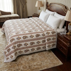 Peut inclure: Un lit est recouvert d'une courtepointe à motifs floraux dans les tons de blanc, marron et or. La courtepointe présente des rayures horizontales et des motifs floraux. Des oreillers blancs reposent contre une tête de lit en bois foncé.