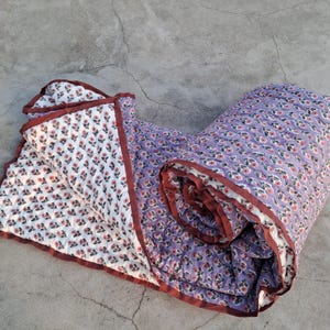Op de afbeelding: Een opgerolde quilt met een bloemenpatroon in wit en paars. De quilt heeft een herhalend patroon van kleine roze bloemen en groene bladeren. Het is afgewerkt met een donkerrode rand.