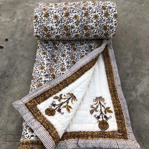 Op de afbeelding: Een opgerolde quilt met een bloemenpatroon in bruin en goud op een witte achtergrond. De quilt is gedeeltelijk uitgerold en toont een wit interieur met bruine bloemmotieven en een gestreepte rand. De quilt is gemaakt van zacht, gewatteerd materiaal.
