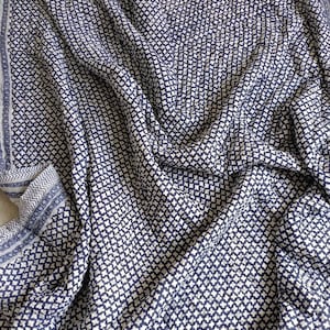 May include: A blue and white patterned textile with a repeating geometric design. The fabric is draped, showing folds and texture. The border features a contrasting pattern. The overall appearance suggests a blanket or throw.