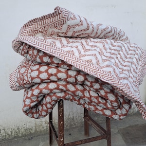 May include: A stack of handmade quilts with various patterns. The top quilt features a white background with a repeating zigzag design in a reddish-brown color. The other quilts have a reddish-brown background with a white leaf pattern.