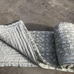 May include: A rolled-up, sage green and white floral patterned quilt with a striped underside. The quilt has a decorative border with a paisley design. The quilt is made of a soft, quilted material.