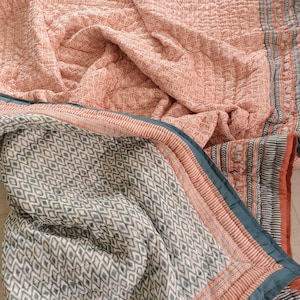 May include: A folded, quilted blanket with a floral pattern in shades of peach and white. The border features stripes and a teal trim. Another blanket with a diamond pattern in teal and white is visible.