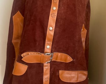 May include: A vintage brown suede coat with tan leather accents. The coat features a round collar, decorative buttons, and a belt with a silver buckle. The coat has two front pockets and leather patches on the shoulders.