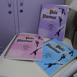 Könnte beinhalten: Drei spiralgebundene Notizbücher mit dem Titel "The Pole Planner" in Lila, Rosa und Blau. Jedes Notizbuch zeigt eine Silhouette einer Pole-Tänzerin und den Text "Angelte Pole Arts". Die Notizbücher liegen auf einer weißen Oberfläche.