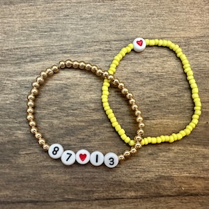 Könnte beinhalten: Zwei Perlenarmbänder, eines mit goldenen Perlen und das andere mit gelben Perlen. Das goldene Armband hat weiße Perlen mit den Zahlen 8, 7, einem Herzen und 13. Das gelbe Armband hat eine weiße Perle mit einem roten Herzen.