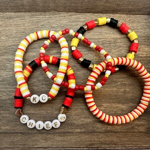 Könnte beinhalten: Ein Set aus vier Armbändern aus roten, weißen und gelben Perlen. Die Armbänder sind übereinander gestapelt und zeigen das Wort "CHIEFS" in weißen Perlen mit goldenen Buchstaben.