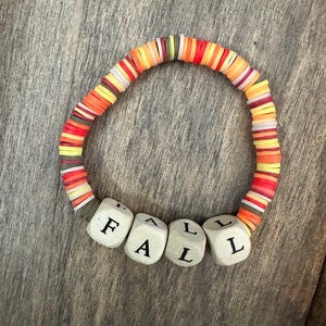 Könnte beinhalten: Ein farbenfrohes Armband zum Thema Herbst mit roten, orangefarbenen und gelben Perlen. Das Armband hat das Wort "FALL" in weißen Perlen geschrieben.