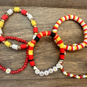 Könnte beinhalten: Satz von fünf Armbändern mit roten, weißen und gelben Perlen. Ein Armband hat das Wort "CHIEFS" in goldenen Buchstaben auf weißen Perlen.