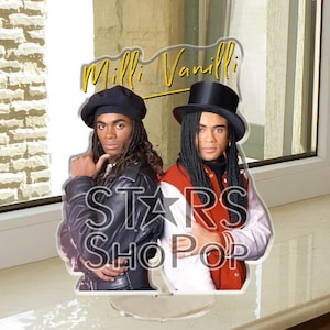 May include: A clear acrylic stand featuring a graphic of Milli Vanilli. The image shows two men, one wearing a black leather jacket and beret, the other in a red and white jacket and top hat. The text "Milli Vanilli" and "Stars Shop Pop" are visible.